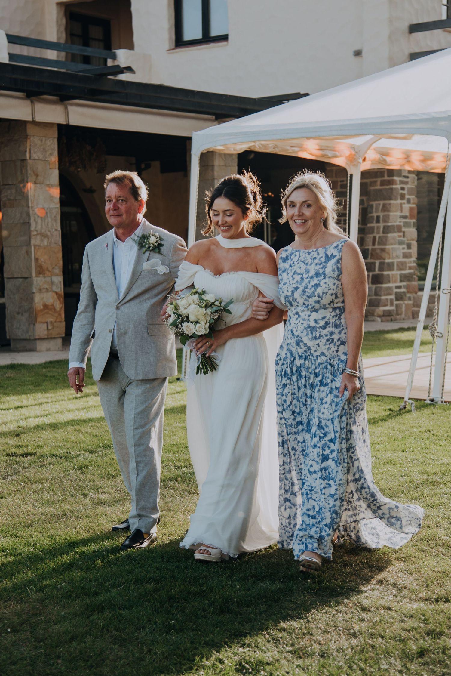 Bride walks down the aisle escorted by parents at outdoor wedding ceremony on green lawn.
