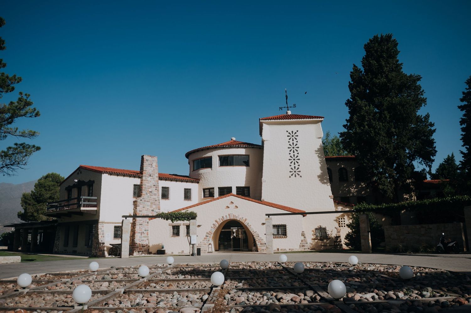 White Spanish-style wedding venue with arched entrance, red tile roof, and tall cypress trees under clear blue sky.
