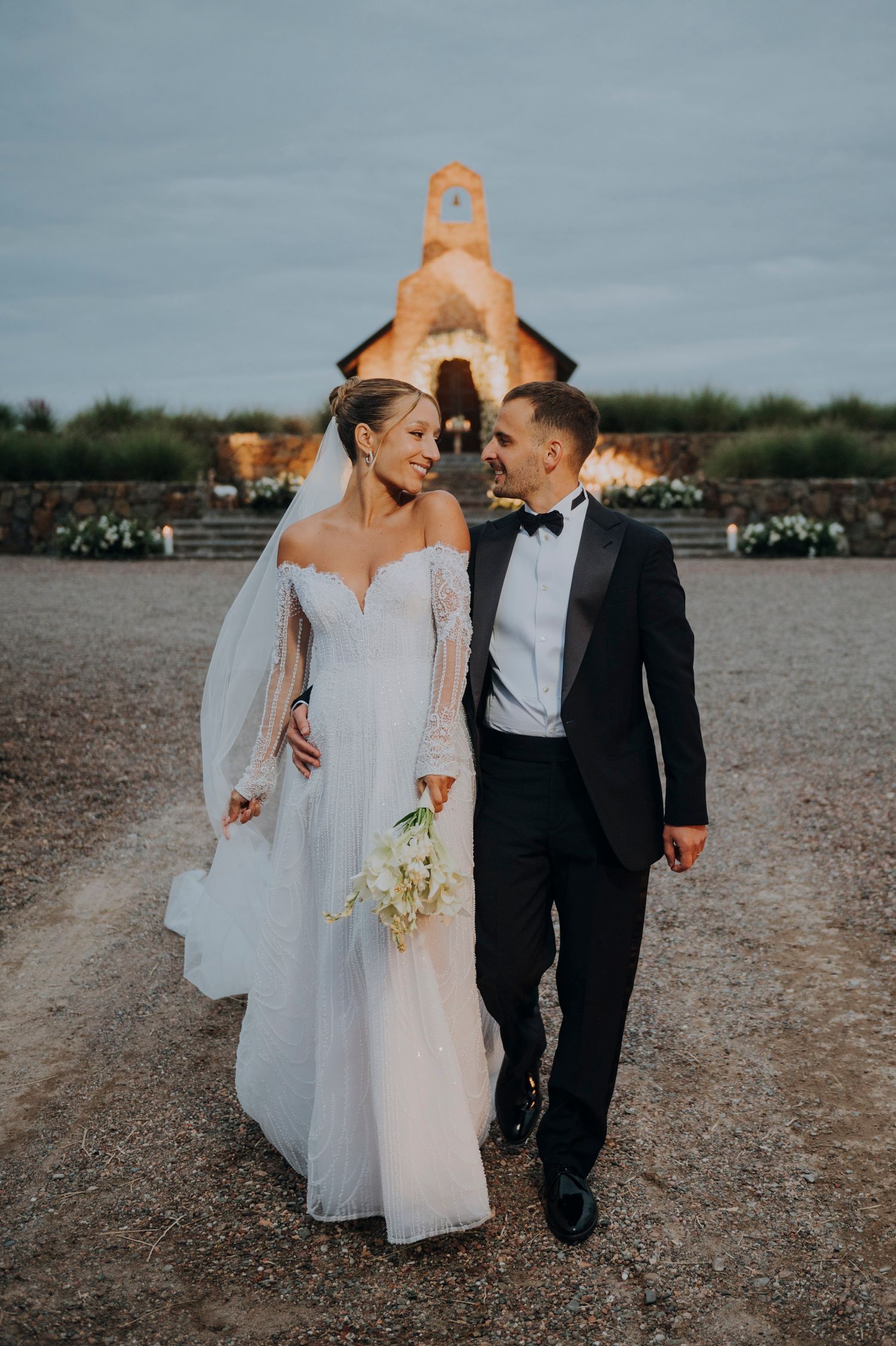 Bride and groom walking together near a rustic stone chapel at dusk. Couple portrait at sunset in Mendoza.
