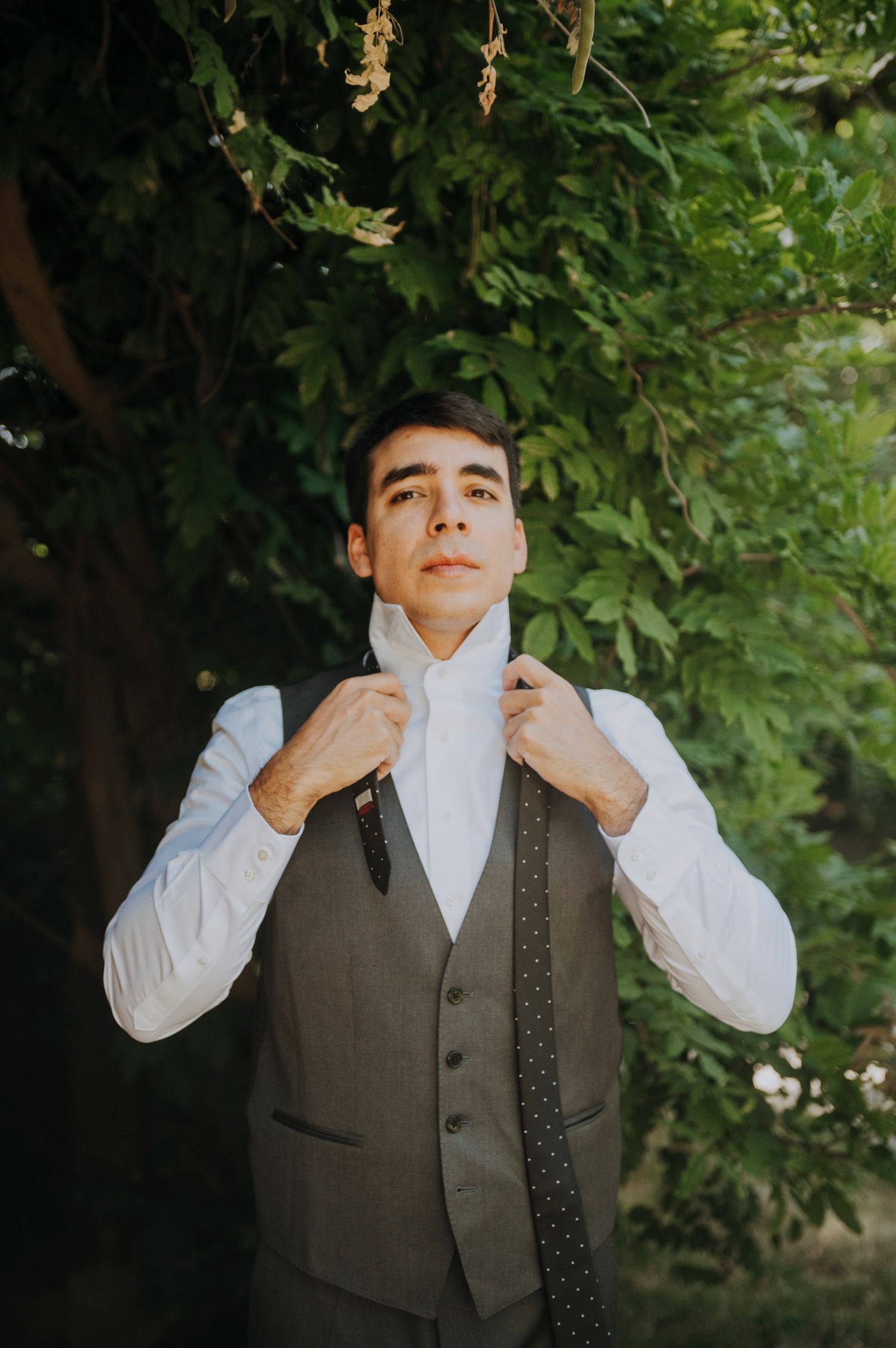 Groom adjusting his tie outdoors wearing a gray vest and white dress shirt before his wedding ceremony.
