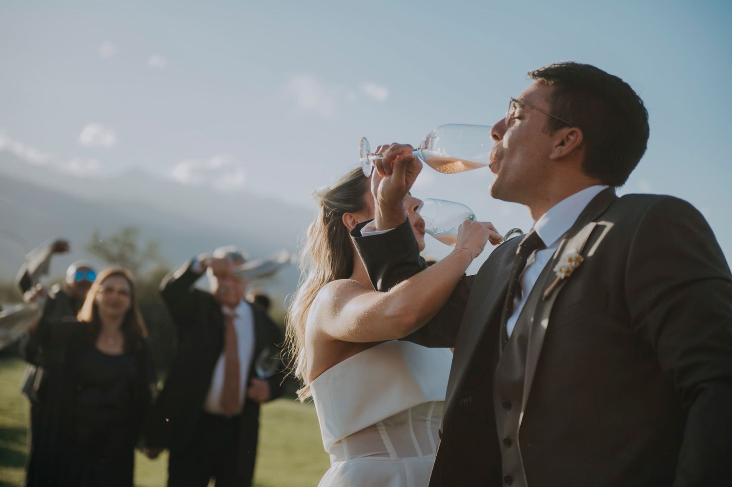 Bride helps groom drink from tilted wine glass at sunny outdoor wedding celebration.