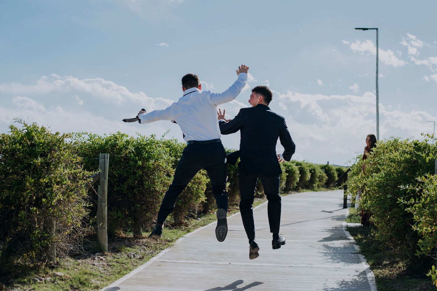 Two groomsmen jumping joyfully on a sunlit garden pathway at a wedding celebration.