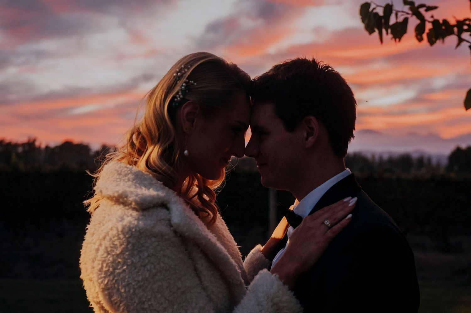 Romantic bride and groom touching foreheads at sunset with dramatic pink and purple sky.