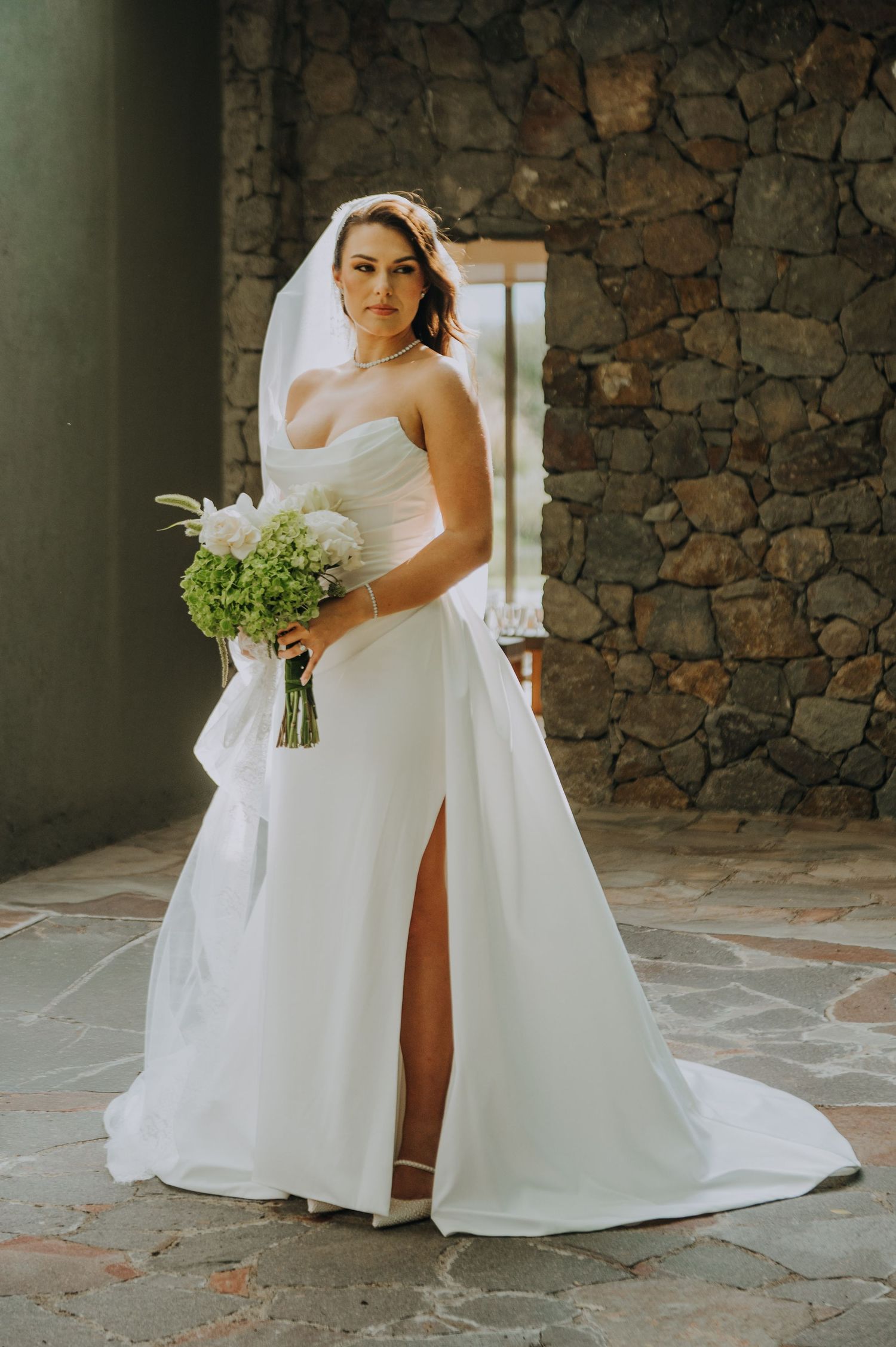 A bride in a strapless white gown with a slit holds a green and white bouquet against a stone wall backdrop.