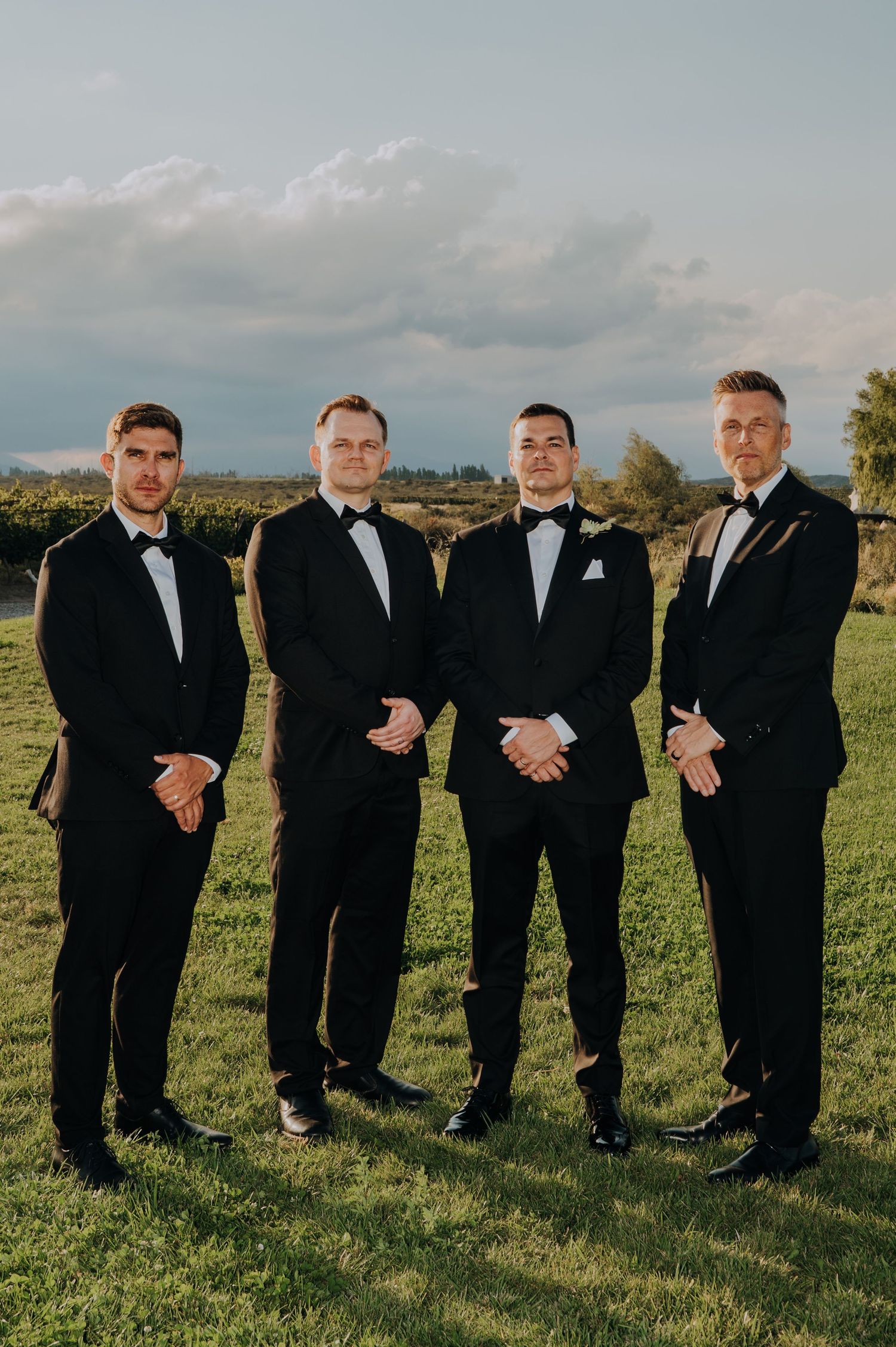 Four groomsmen in black tuxedos with bow ties posing outdoors on green grass with a cloudy sky backdrop.