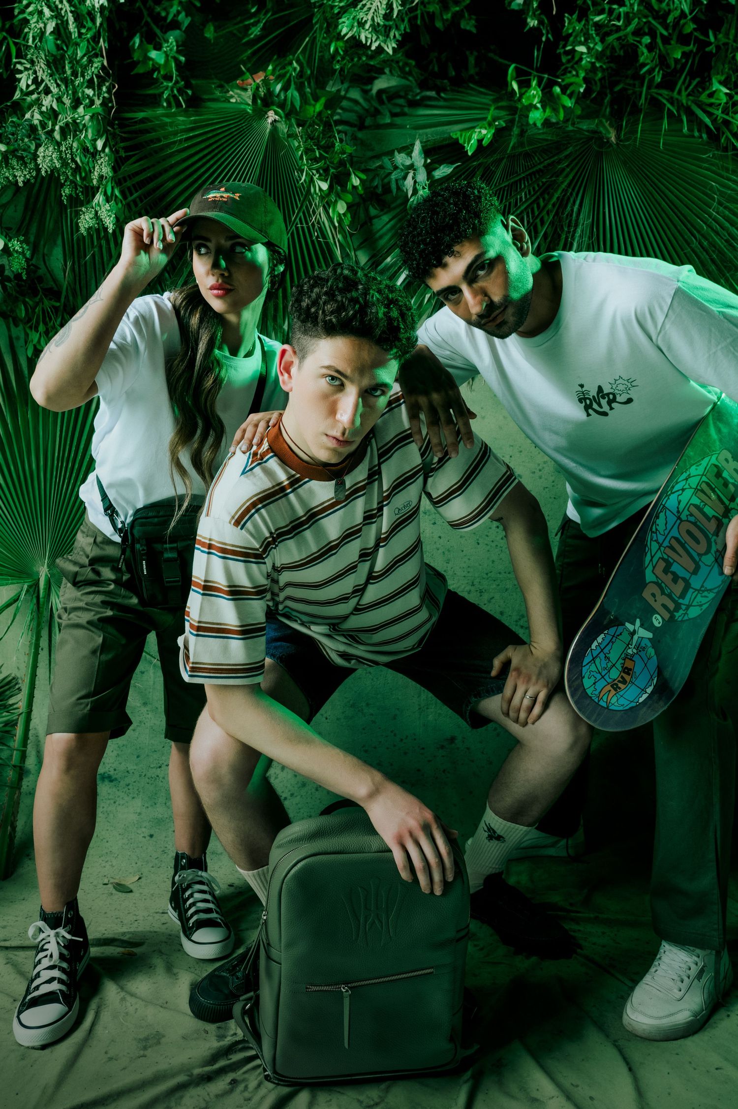 Three young friends pose together in green-lit tropical setting with skateboard and backpack.