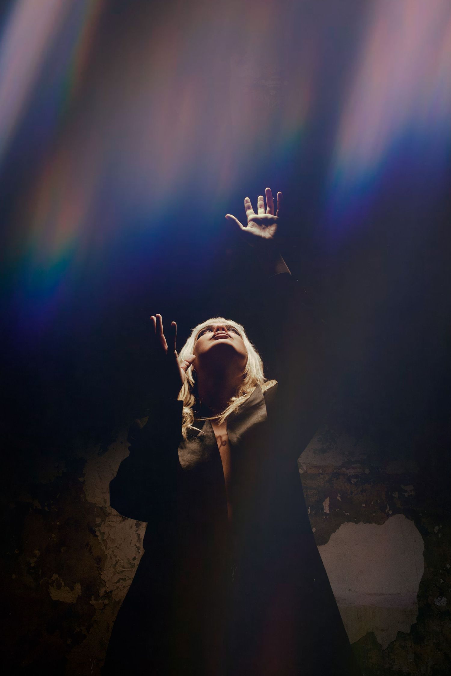 MORA NAVARRO Blonde person in dark coat reaching upward toward colorful rainbow light rays in dark setting.