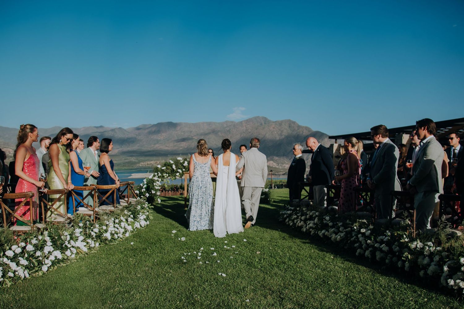 Outdoor wedding ceremony on green lawn with mountain backdrop and guests lining the aisle.