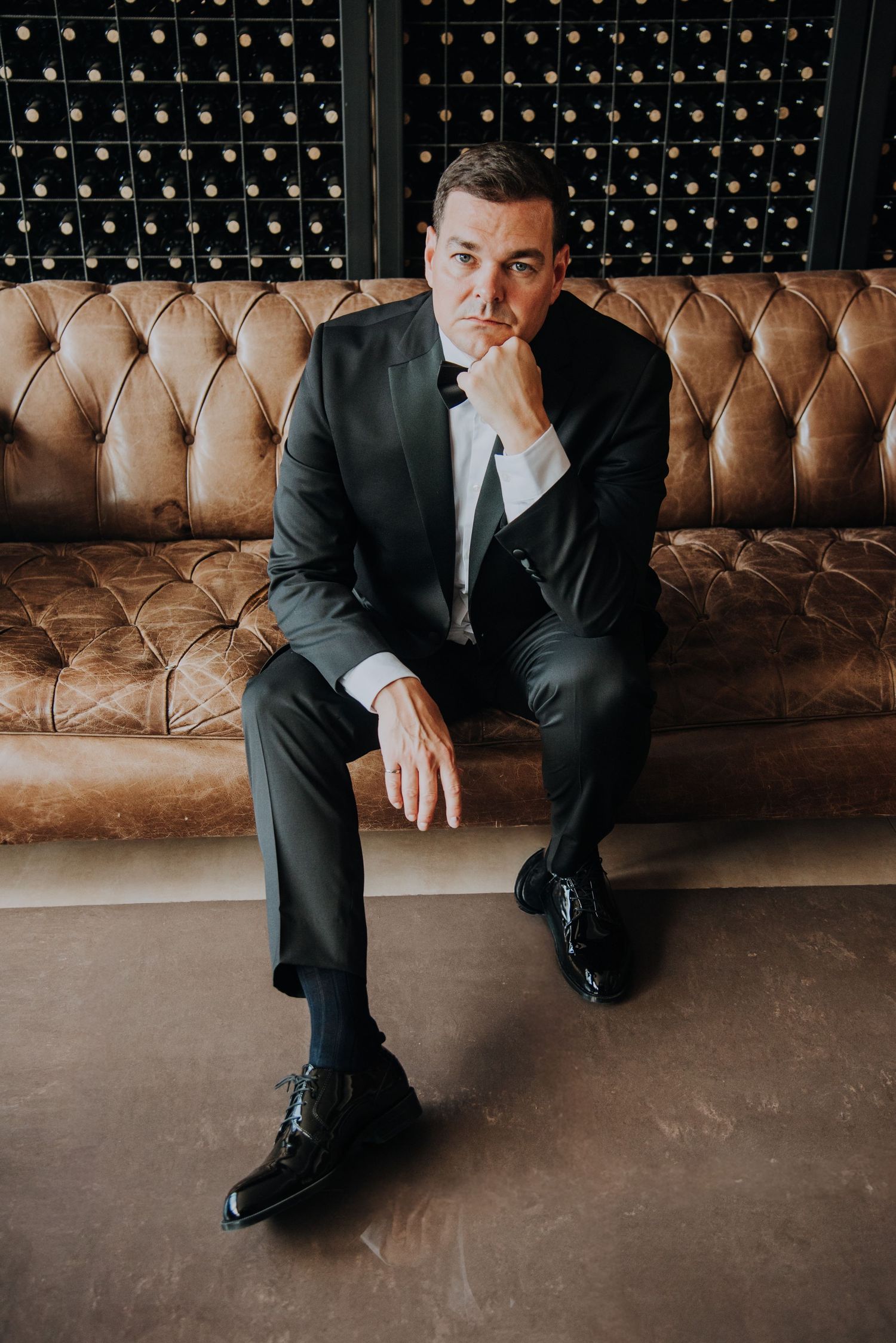 Elegant groom in dark suit sitting on tufted leather sofa in a wine cellar.