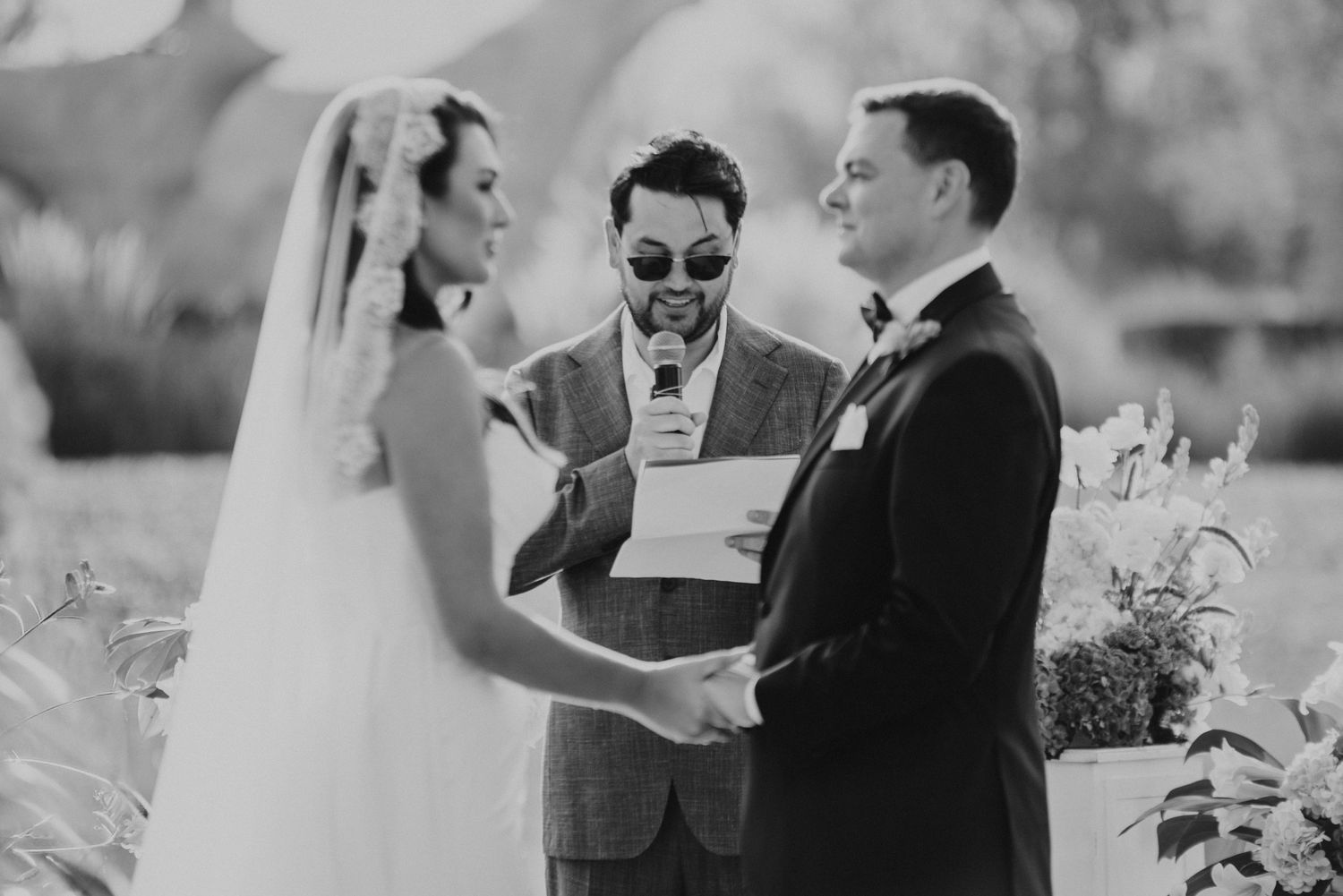 Black and white photo of bride and groom exchanging vows with officiant at outdoor wedding ceremony.