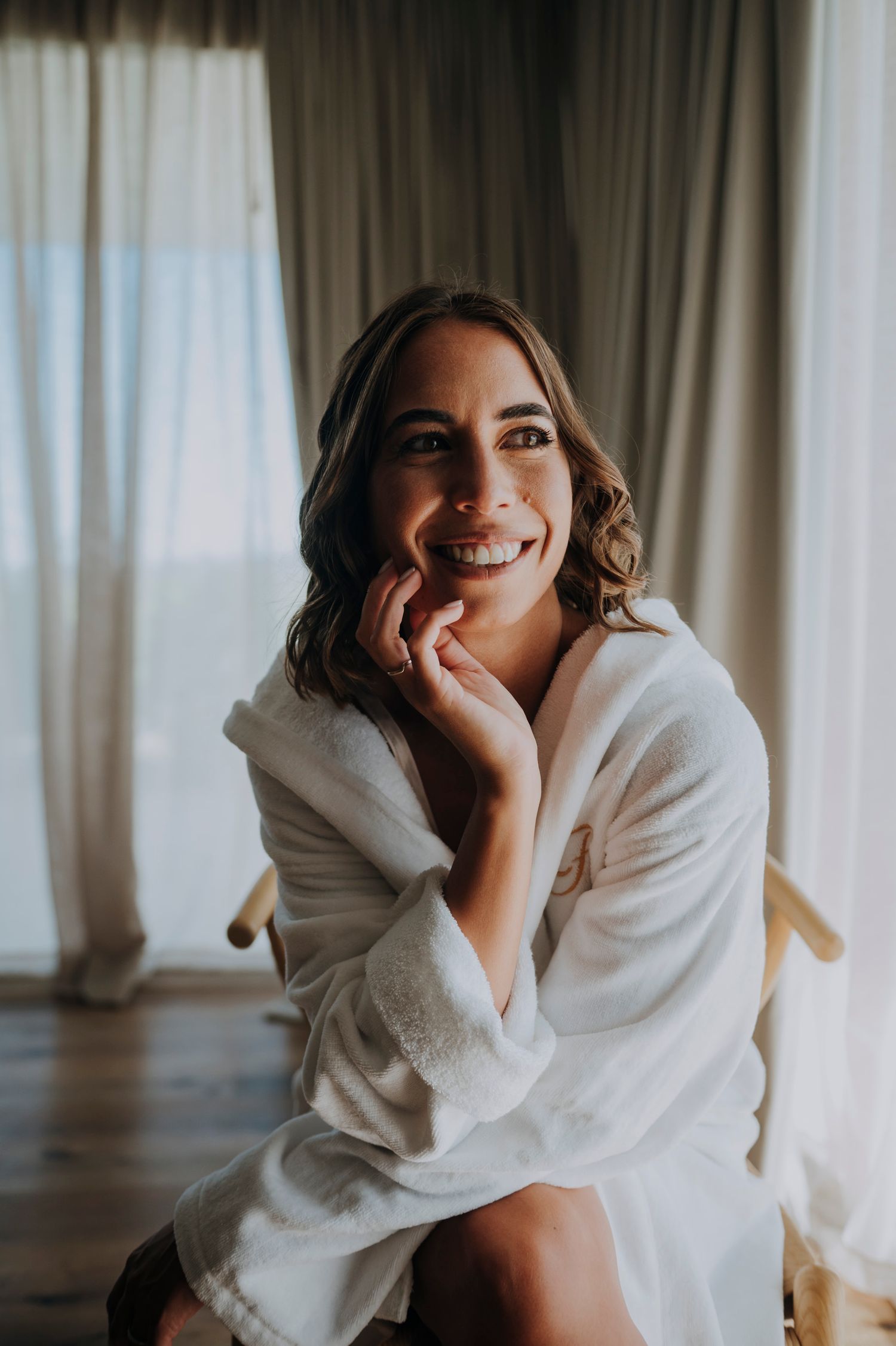 Radiant bride in white robe sitting and smiling warmly during wedding morning preparations.