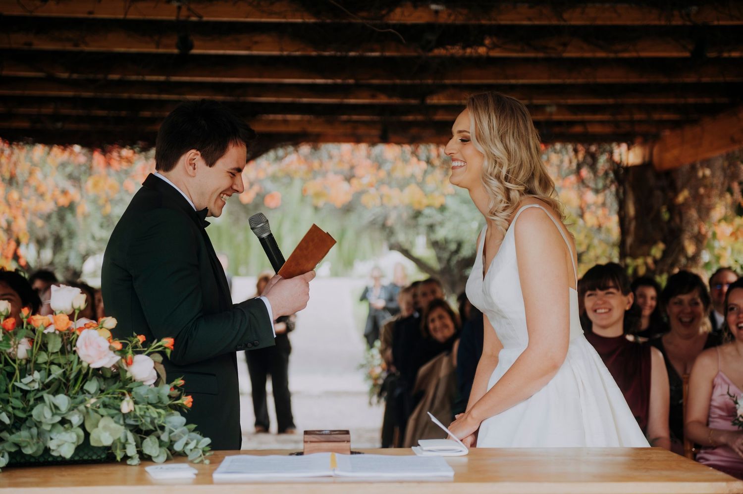 Groom reads heartfelt vows to smiling bride at elegant outdoor wedding ceremony altar with floral arrangements.