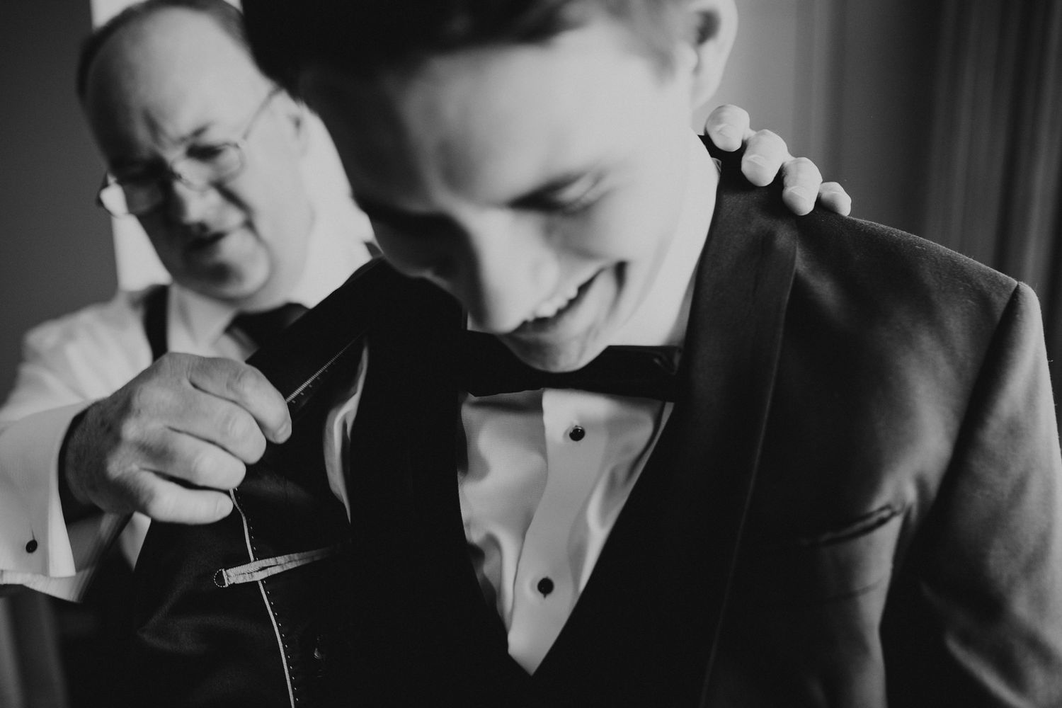 Black and white photo of a groom smiling as an older man helps pin a boutonniere on his tuxedo lapel.