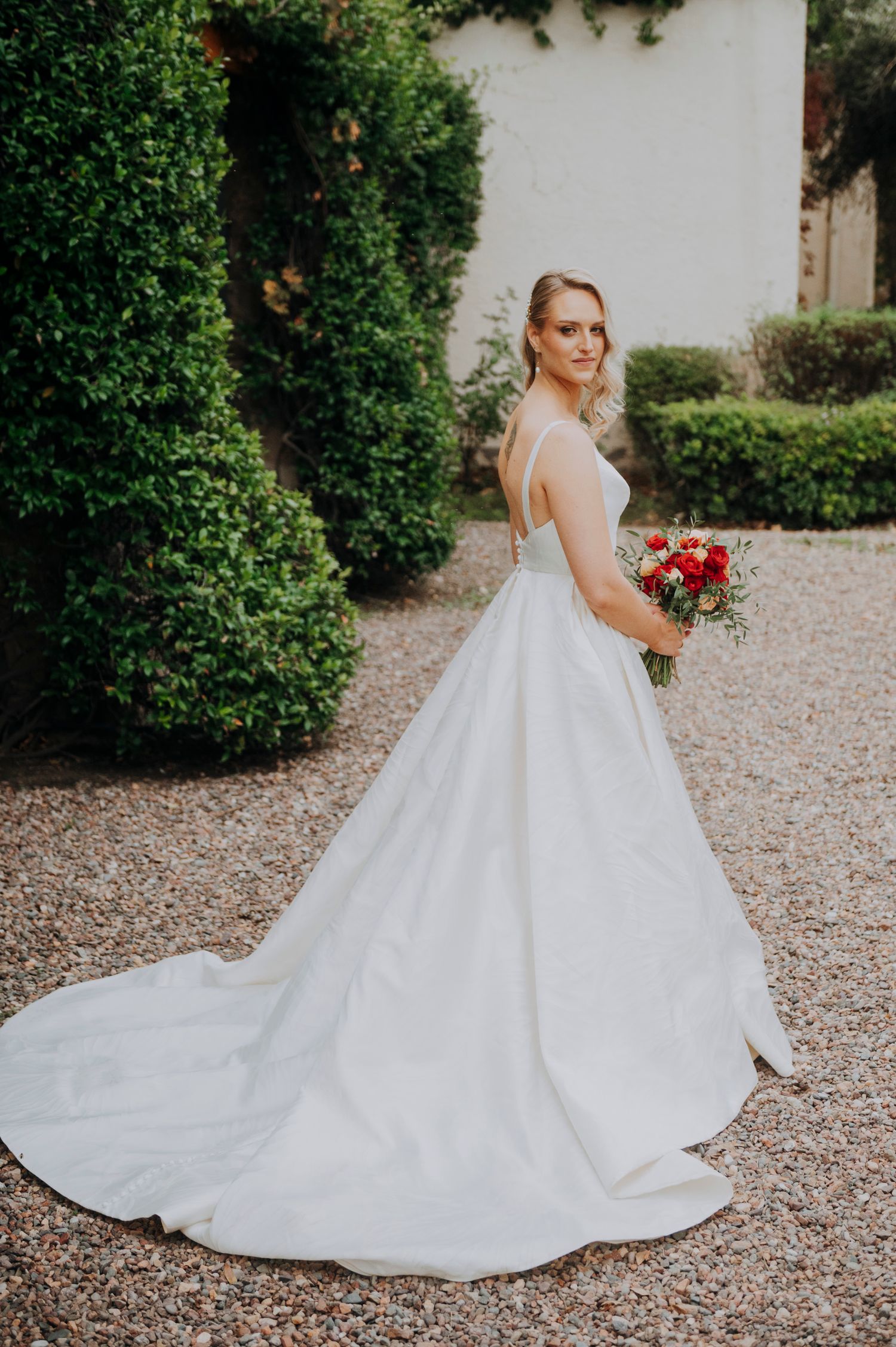 Bride in elegant white ballgown with long train holds red bouquet in manicured garden setting.