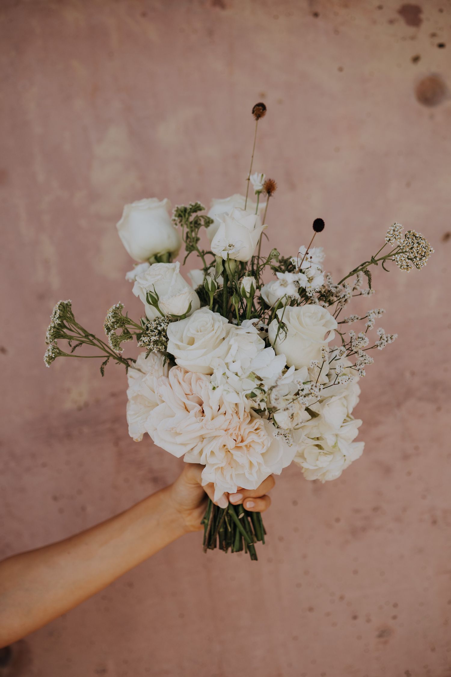 Bride holding elegant white wedding bouquet with roses and wildflowers against pink wall.