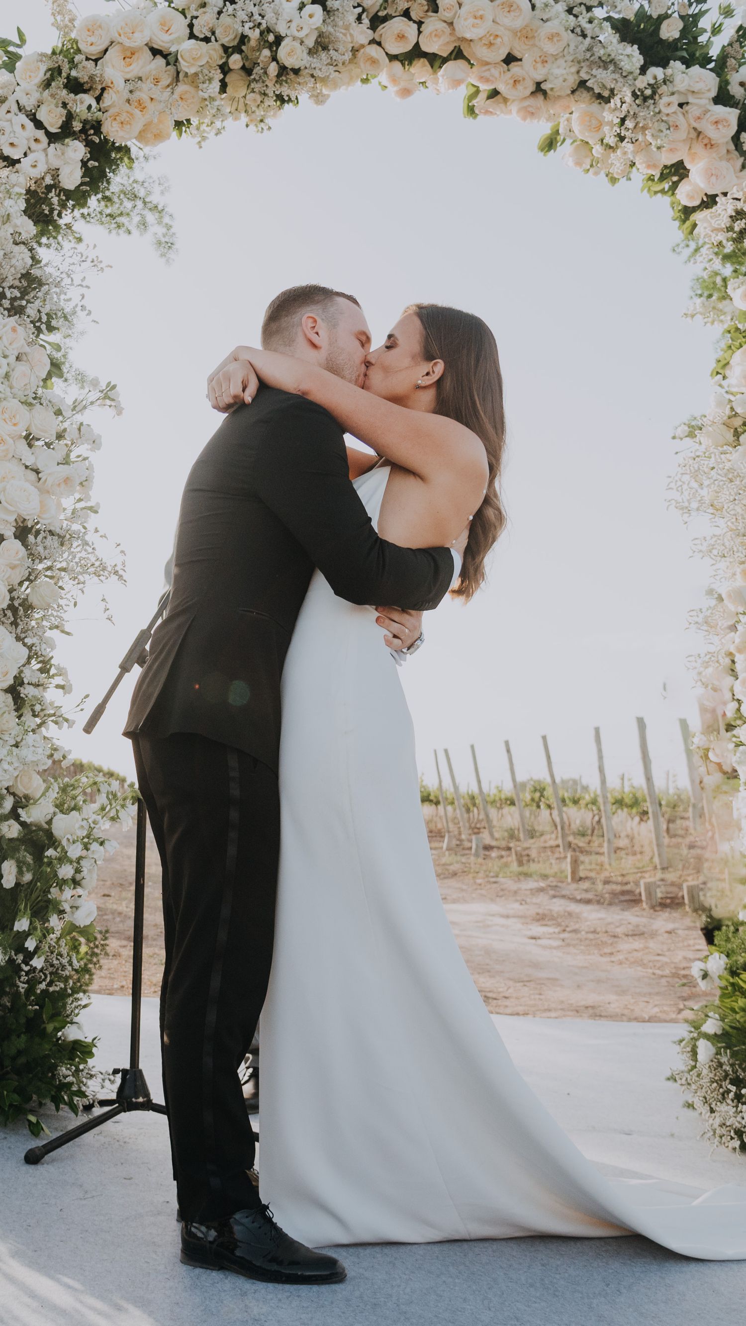 Bride and groom share first kiss under stunning white floral arch at outdoor vineyard wedding ceremony.
