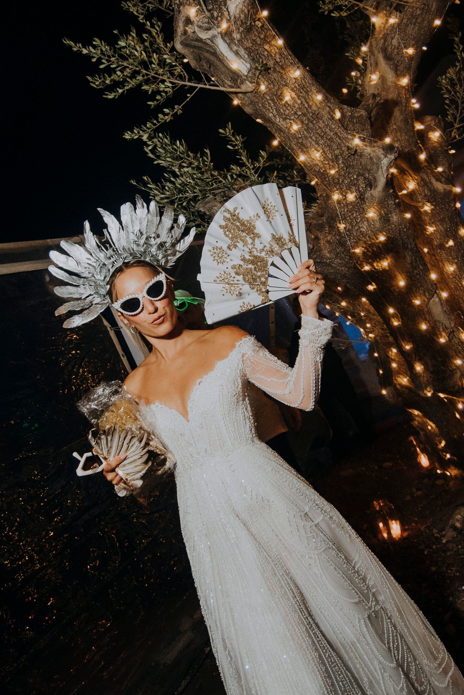 Bride poses in a sparkling gown with feather crown, white fan, and sunglasses under fairy lights.