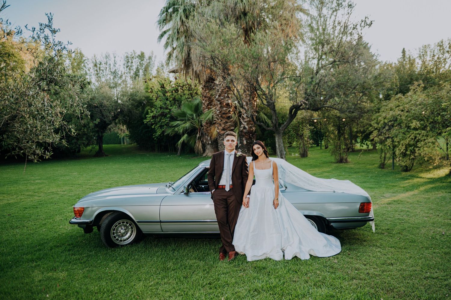 Bride and groom posing beside a vintage silver convertible car in a lush green garden on their wedding day.