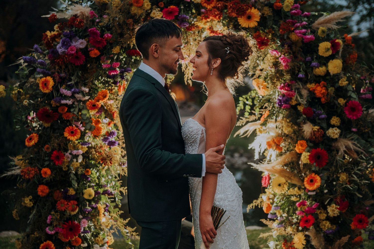 Romantic couple nearly kissing under vibrant floral wedding arch at sunset.Couple portrait at sunset in Mendoza.