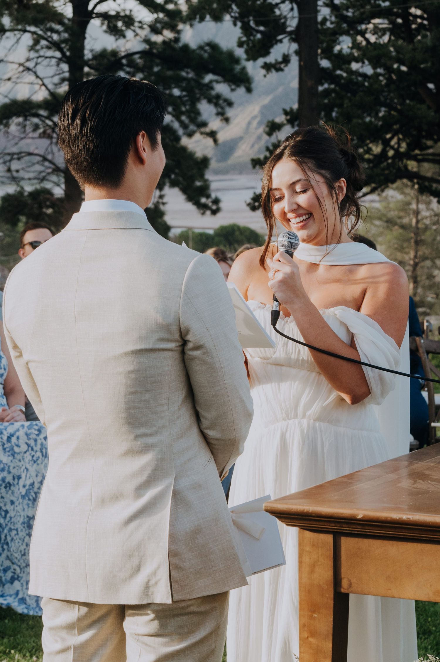 Bride smiling and reading vows into microphone at outdoor wedding ceremony with groom in beige suit.