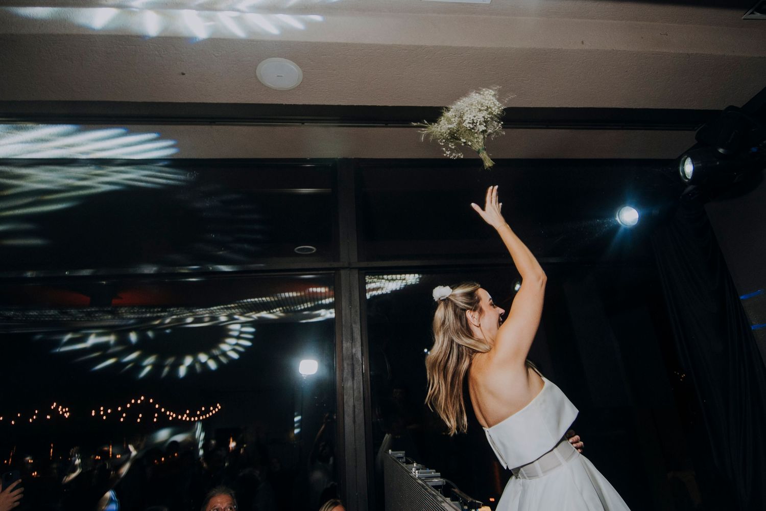 Bride in white strapless gown tosses her bouquet to guests during indoor wedding reception celebration.