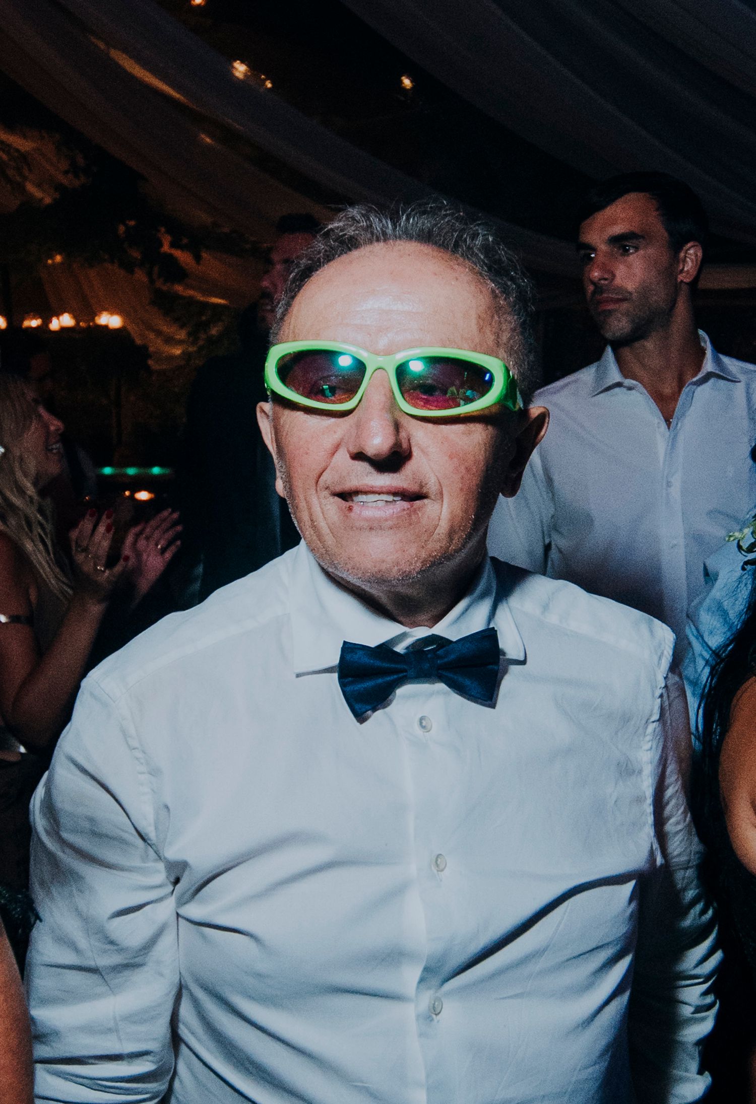 Cheerful wedding guest wearing neon green sunglasses and blue bow tie dancing at a reception.