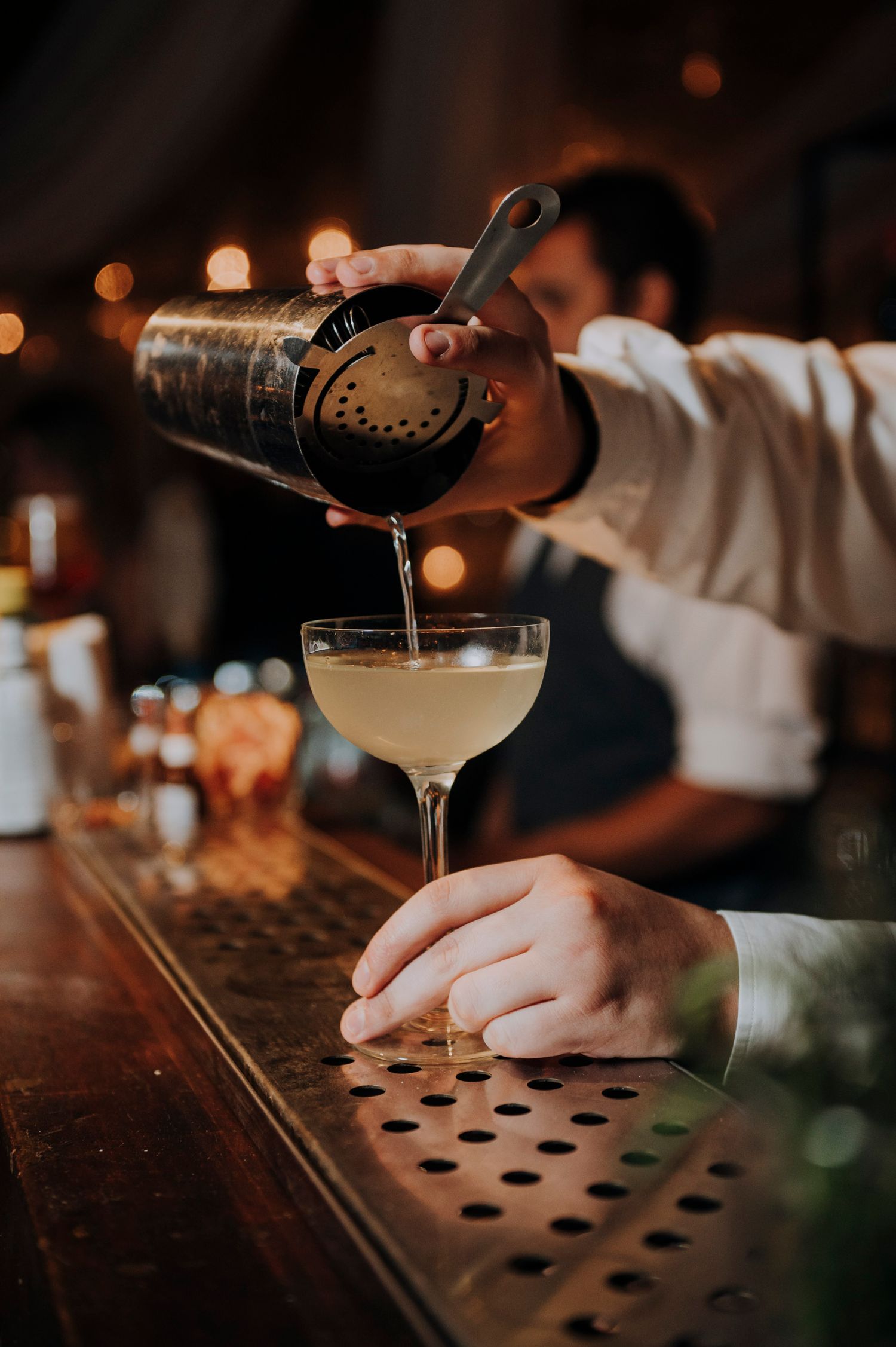 Bartender pouring a cocktail through a strainer into a coupe glass at a wedding bar.