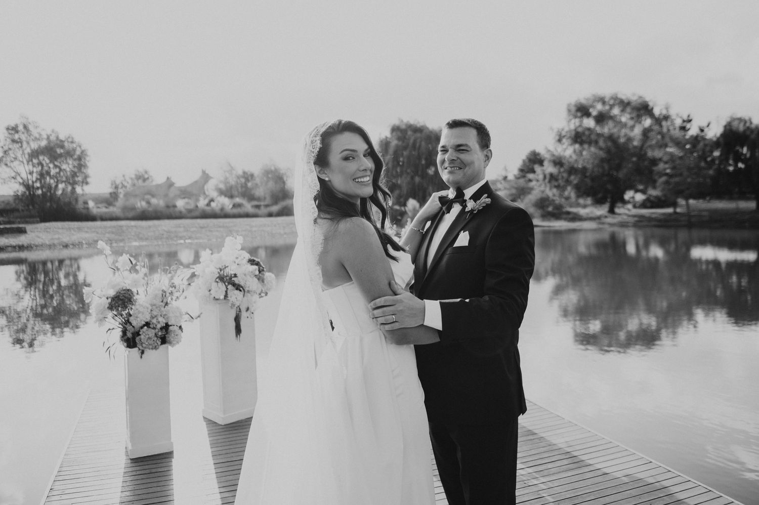 Black and white portrait of bride and groom embracing on a dock with a serene lake backdrop.