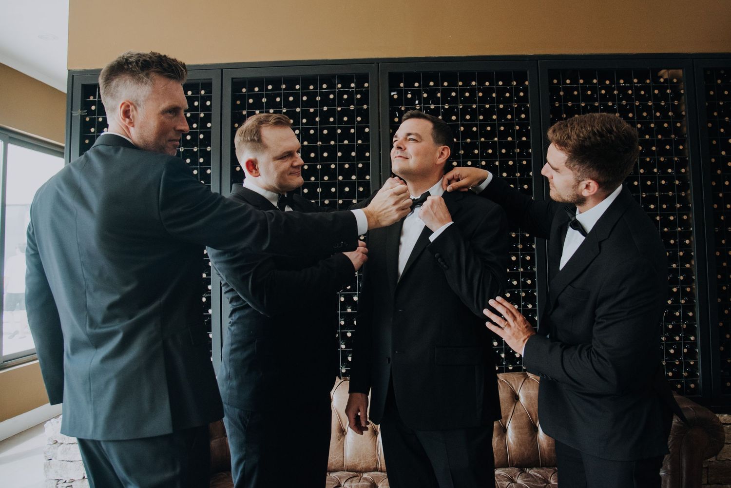 Groomsmen in black suits helping groom adjust his tie before wedding ceremony indoors.