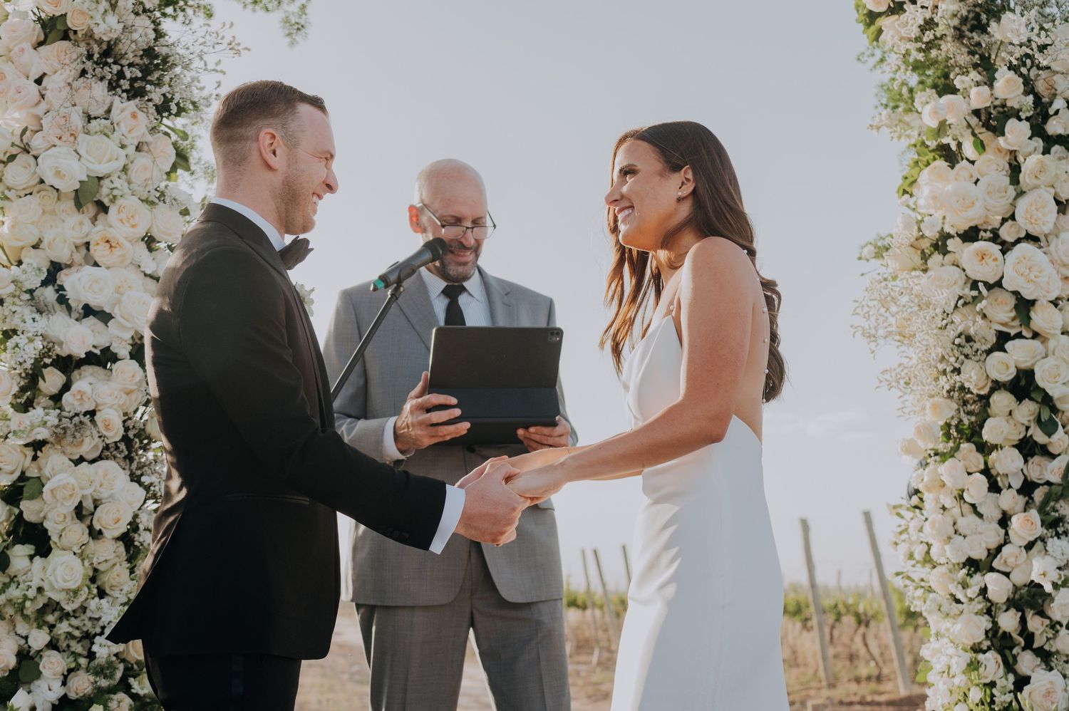 Couple exchanging vows at outdoor wedding ceremony with white floral arch and officiant.