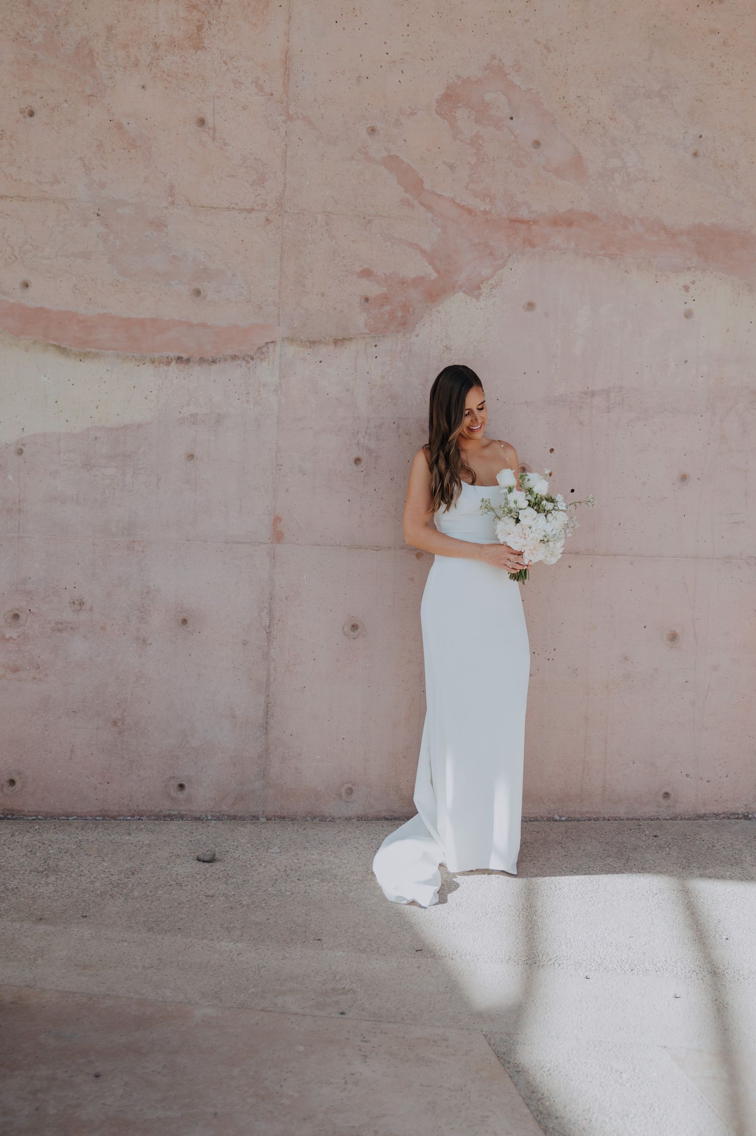 Bride in elegant white gown holding white bouquet, standing against a pink concrete wall in soft natural light.