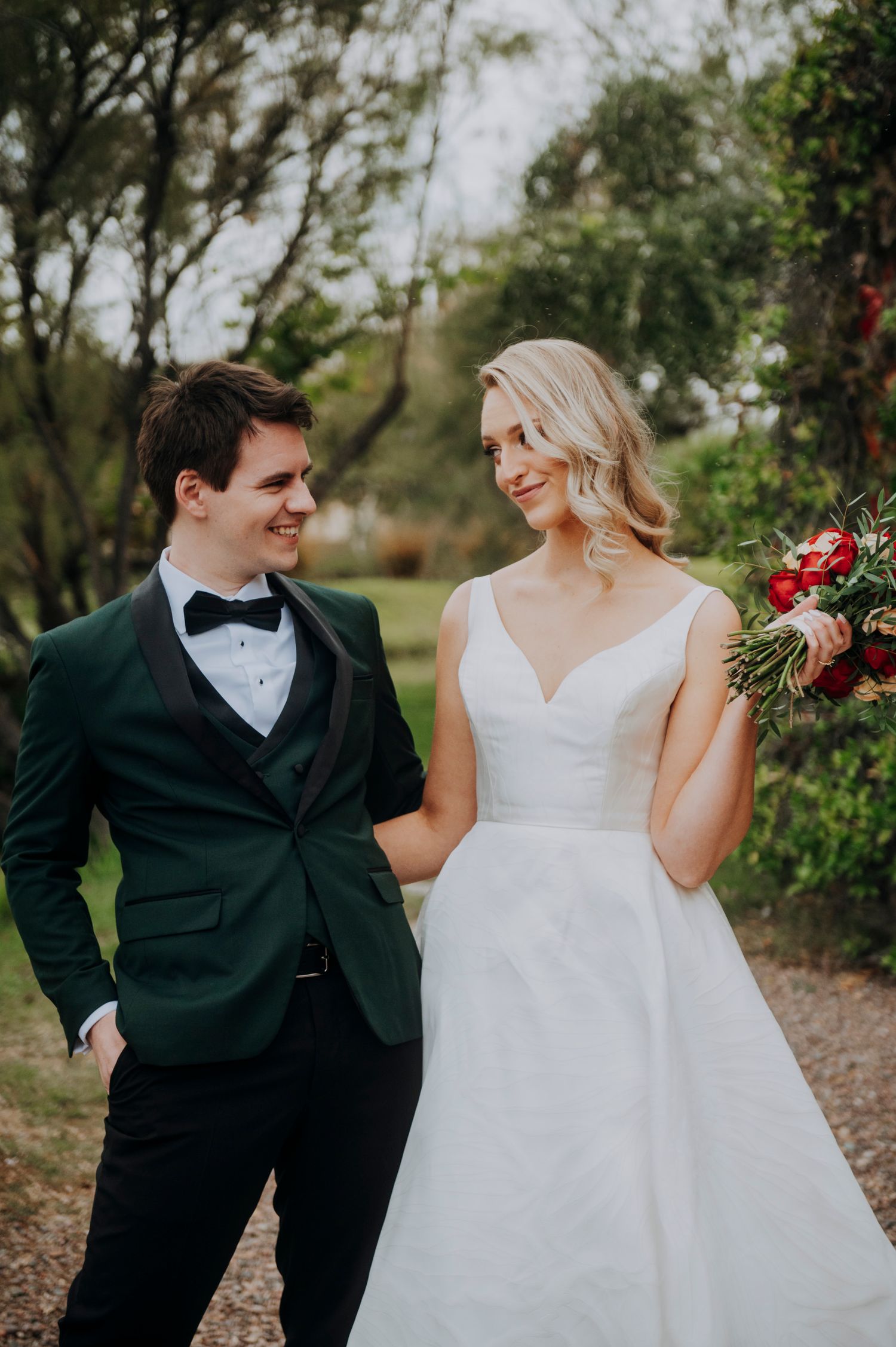 Bride and groom walking together outdoors, she holds a red bouquet wearing a white gown, he wears a green tuxedo.