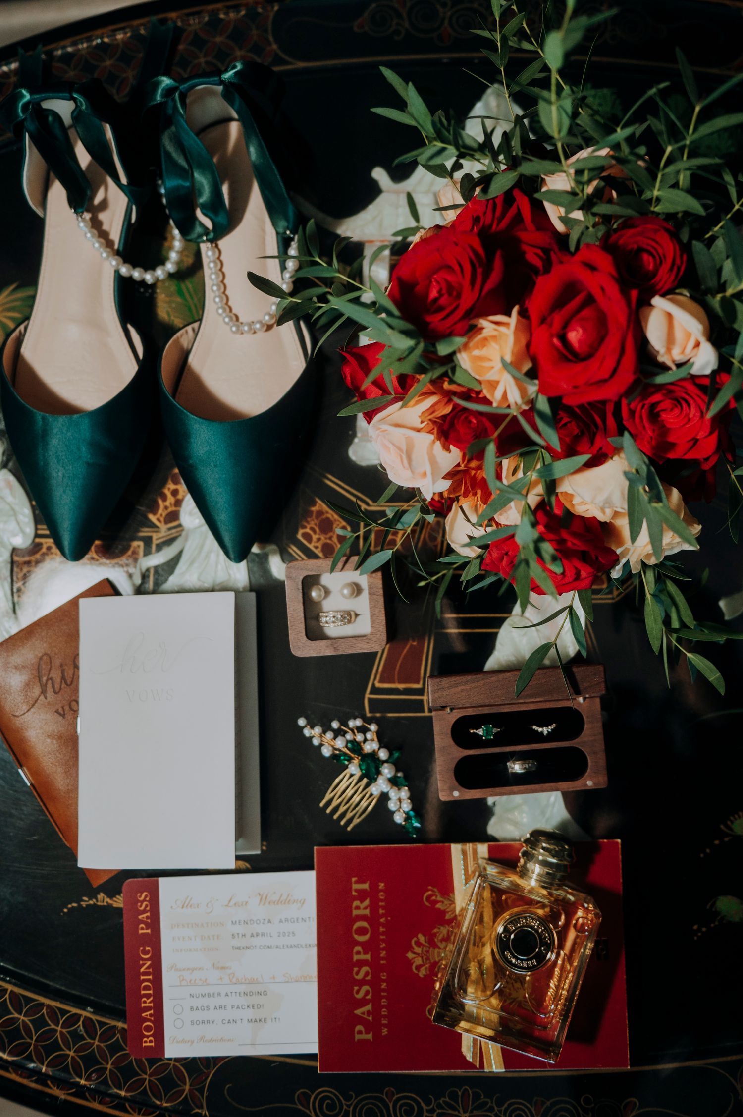 Elegant wedding flat lay with teal heels, red rose bouquet, passport invitation, rings, and gold perfume on dark surface.