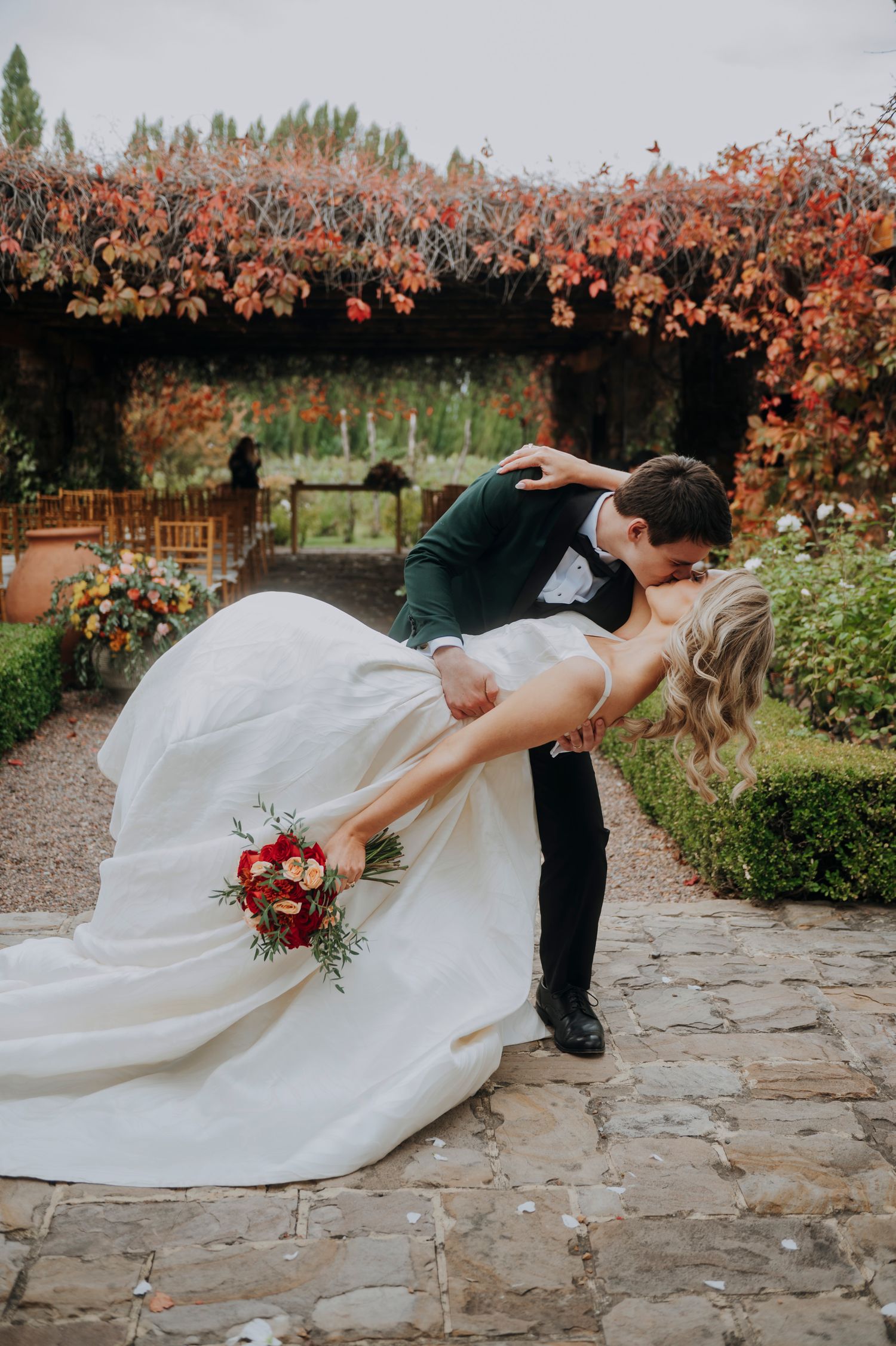 Groom dips bride in white gown holding red bouquet, surrounded by autumn foliage at outdoor wedding venue.