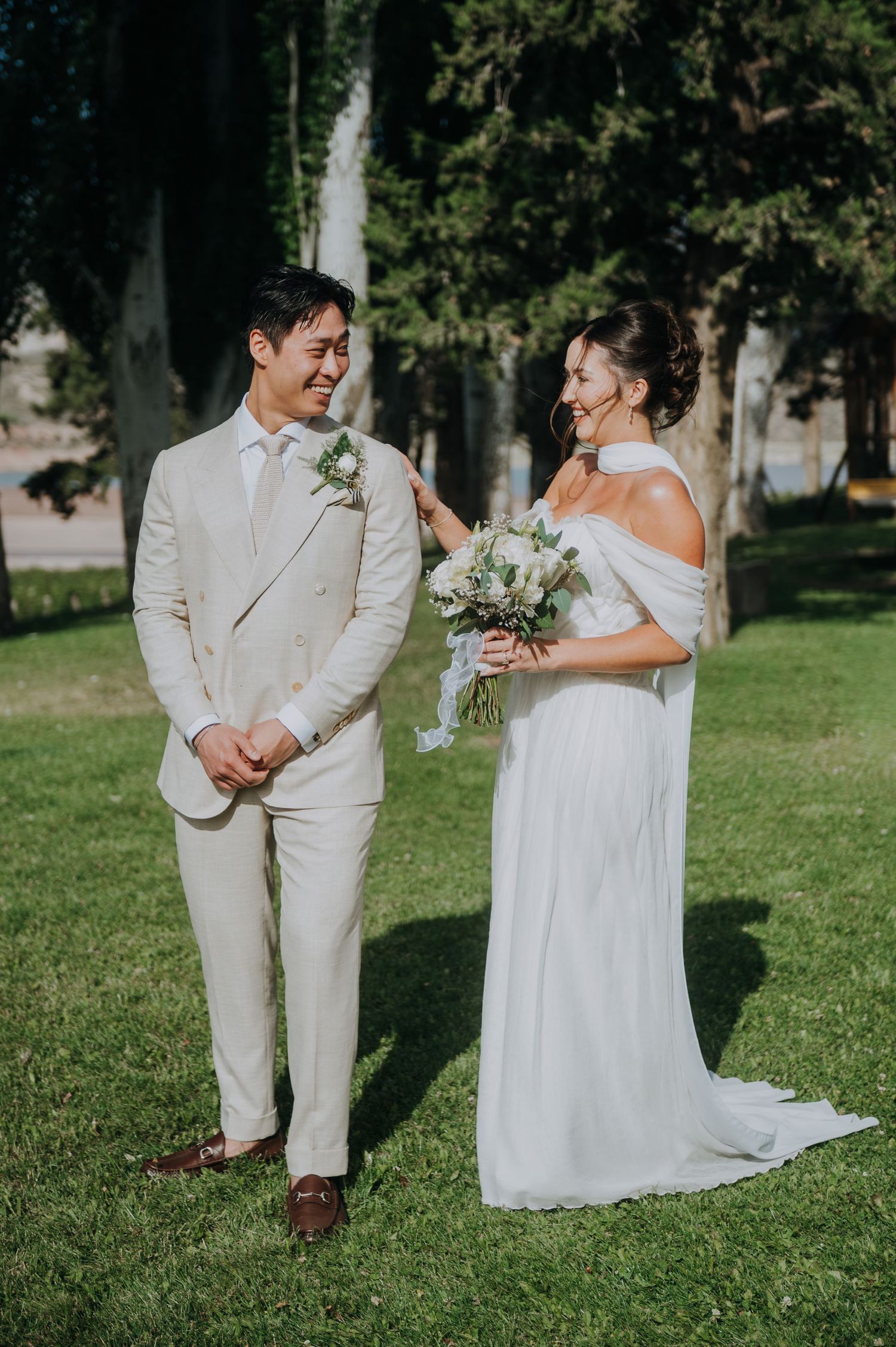Smiling groom in beige suit and bride in white off-shoulder gown holding bouquet during outdoor wedding first look.