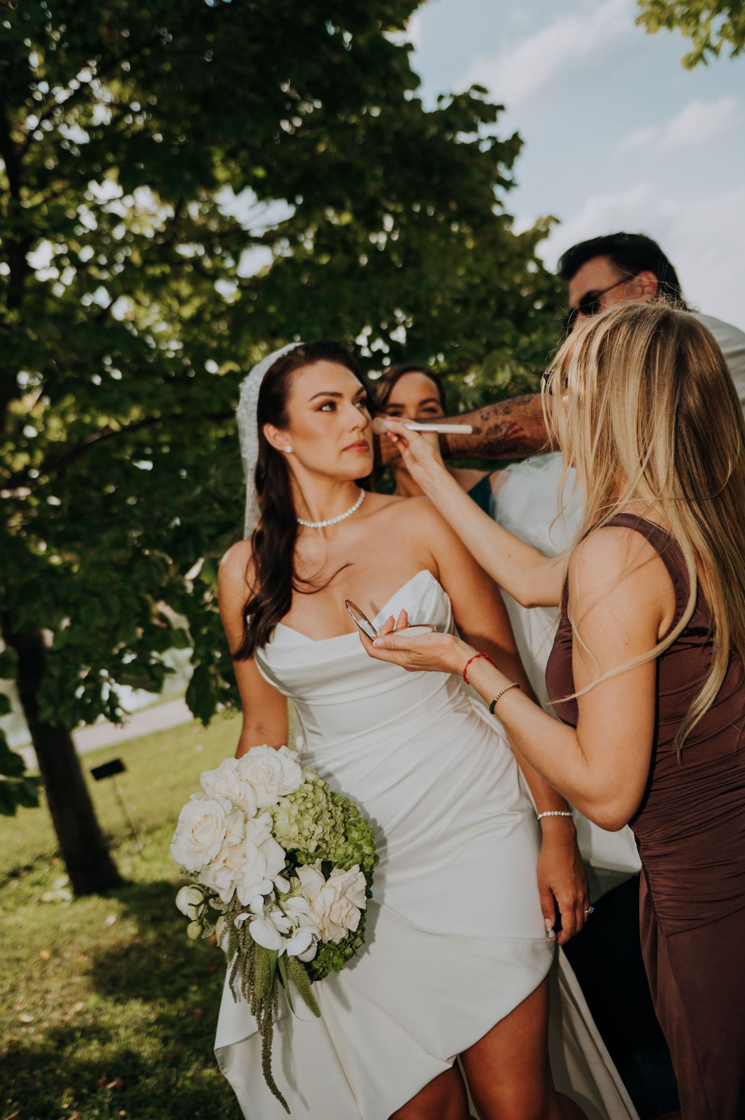 Bridesmaid touching up bride's makeup outdoors before wedding ceremony, bride holding white bouquet.