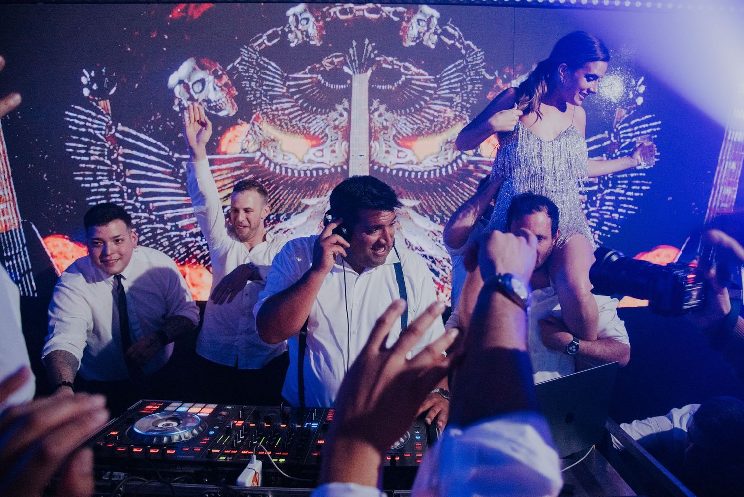 Lively wedding reception with a DJ and guests dancing under blue lights and a dramatic stage backdrop.