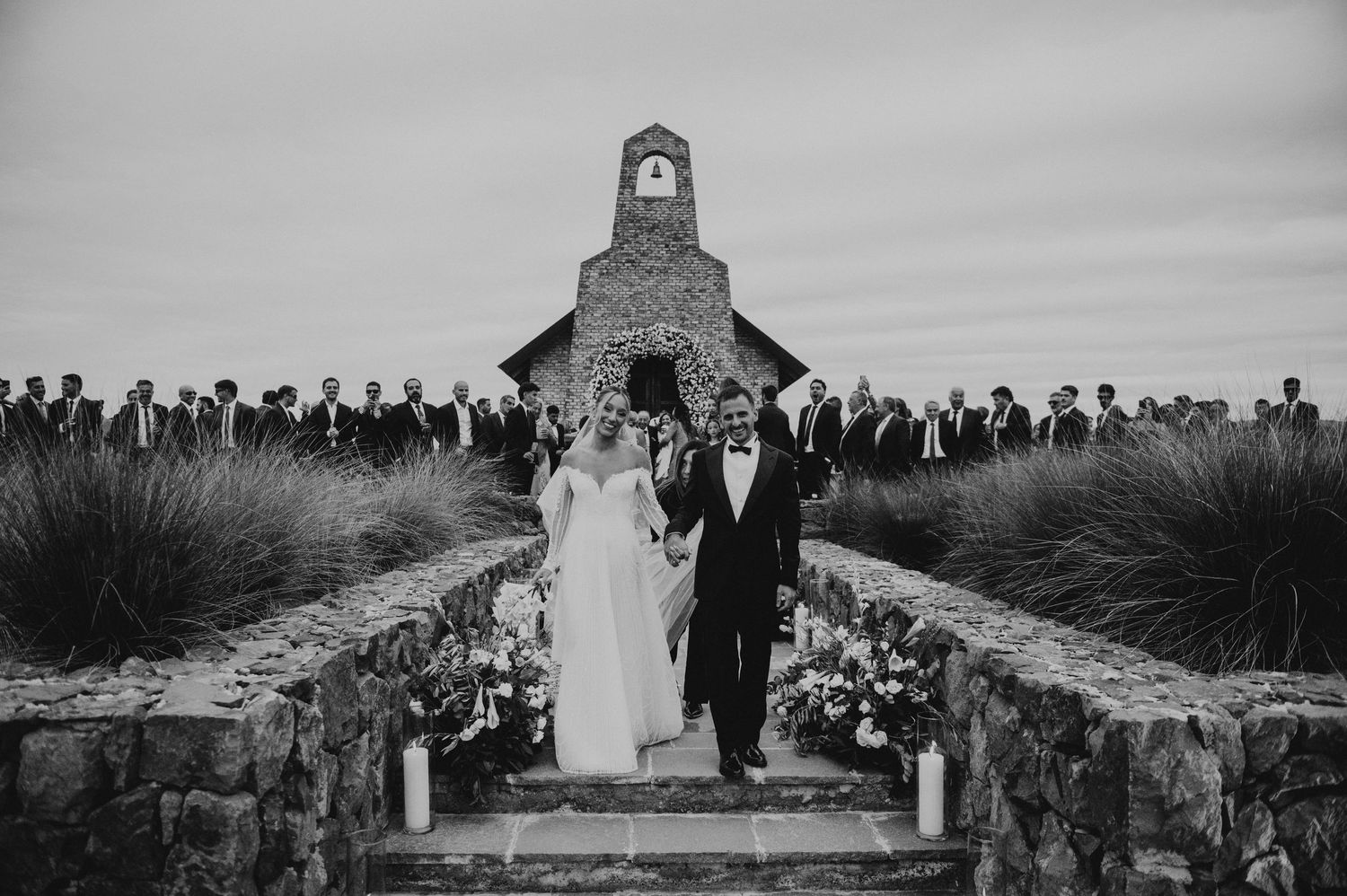 Black and white wedding photo of bride and groom exiting stone chapel surrounded by guests in scenic outdoor setting.
