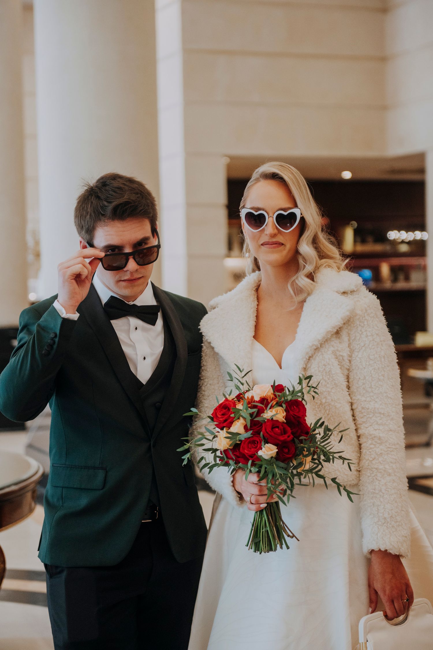 Bride and groom in green suit wear sunglasses holding red rose bouquet in elegant wedding venue.