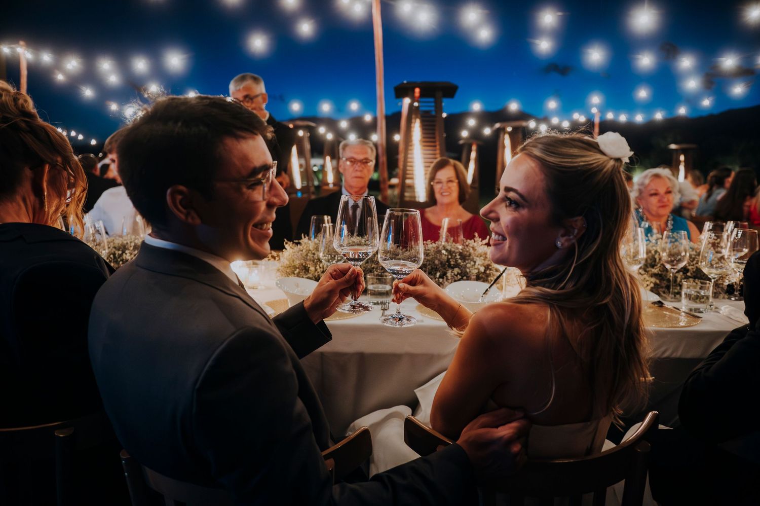 Bride and groom toast with wine glasses at elegantly lit outdoor evening wedding reception.