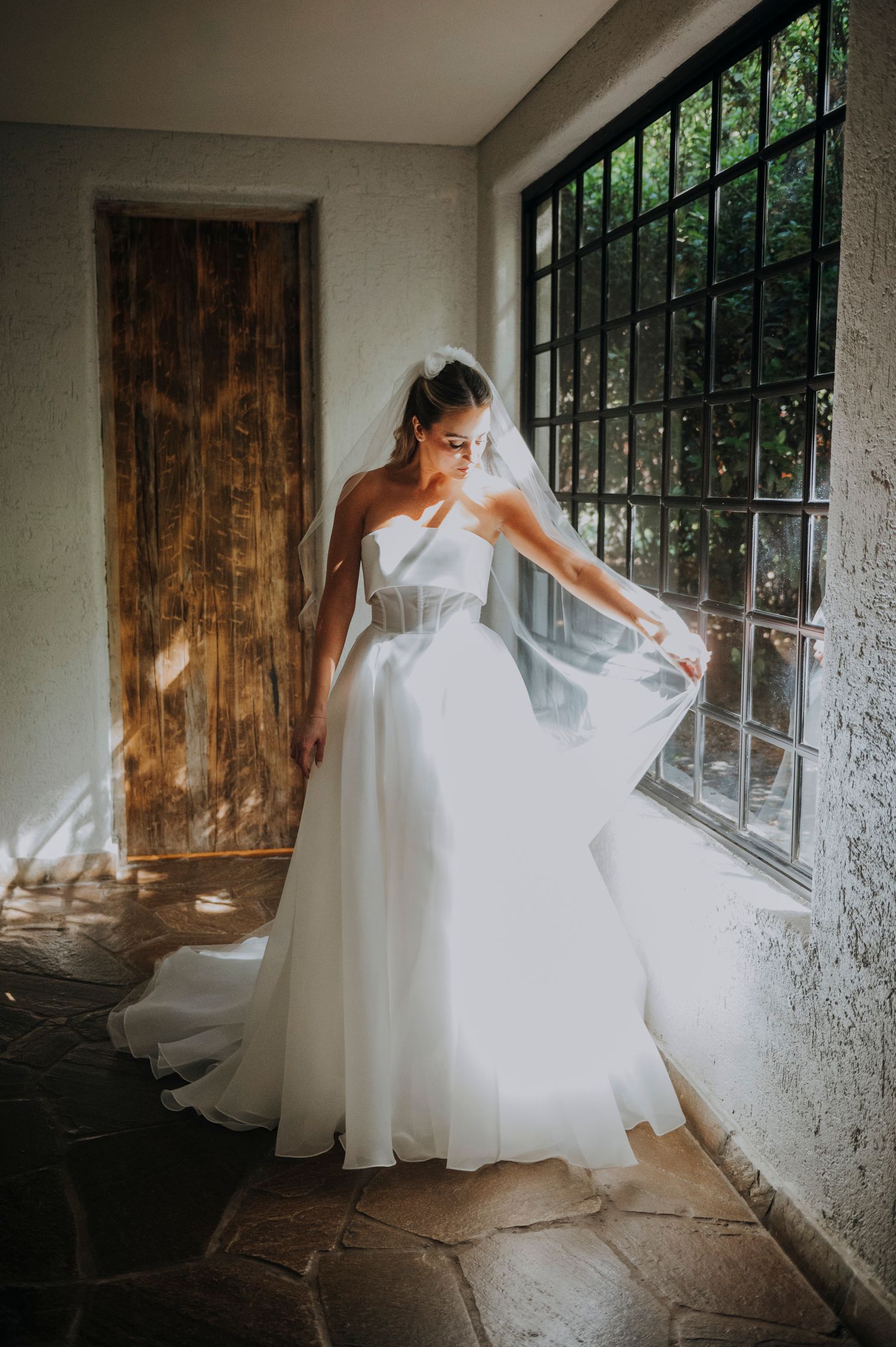 Bride in white strapless ballgown and veil posing by window in rustic venue.