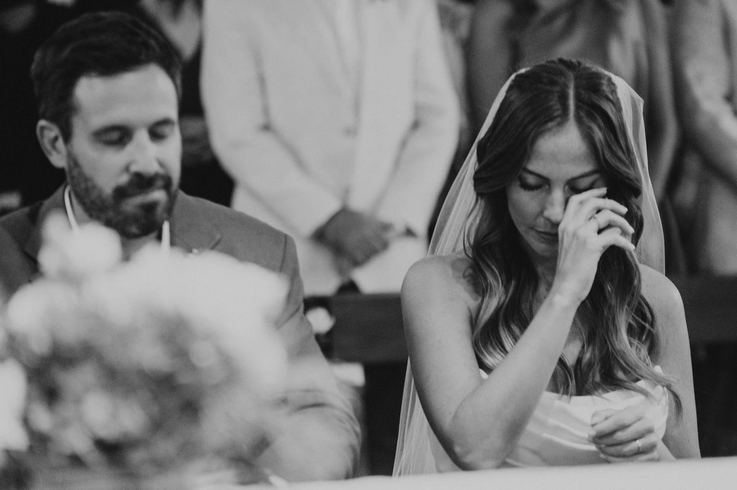 Emotional bride in veil praying at wedding ceremony, groom beside her, black and white photo.