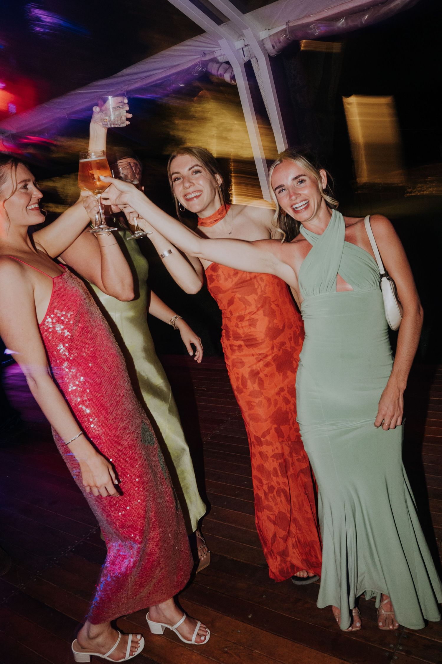 Four women in elegant gowns toasting drinks at a festive evening party celebration.