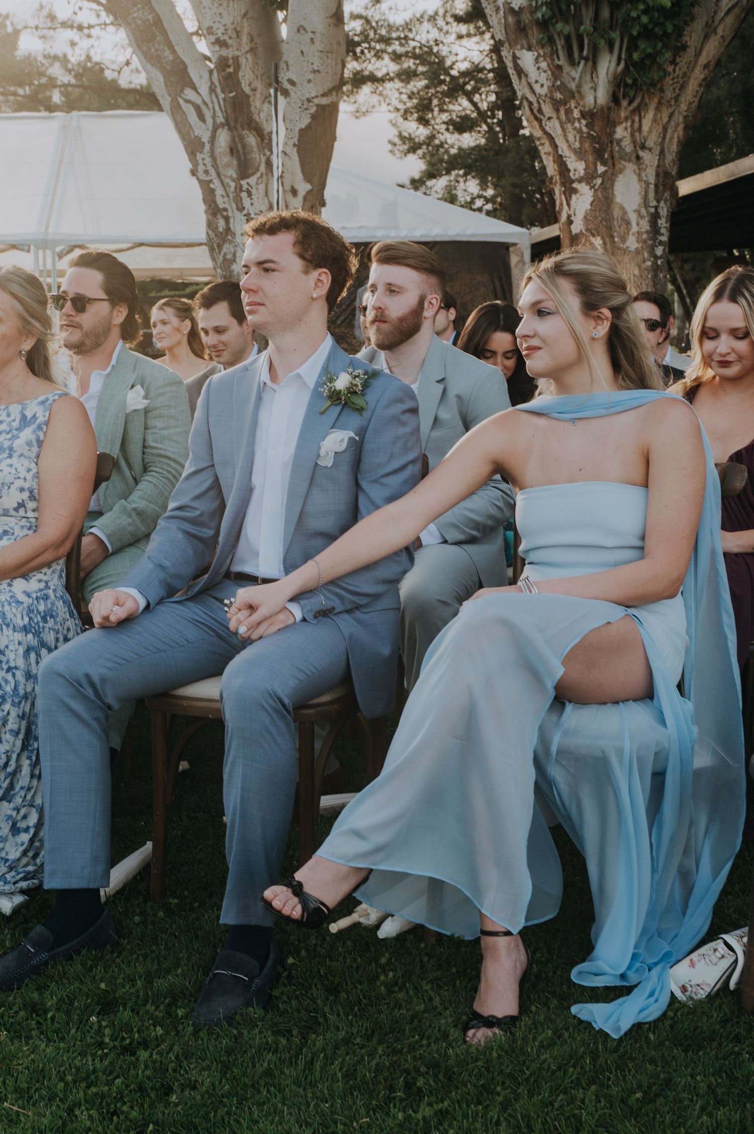 Wedding guests seated outdoors, man in grey suit beside woman in light blue strapless gown.
