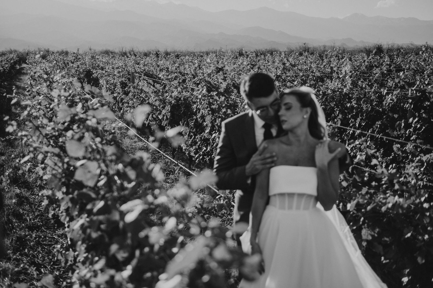 Black and white romantic photo of bride and groom embracing in a lush vineyard setting.