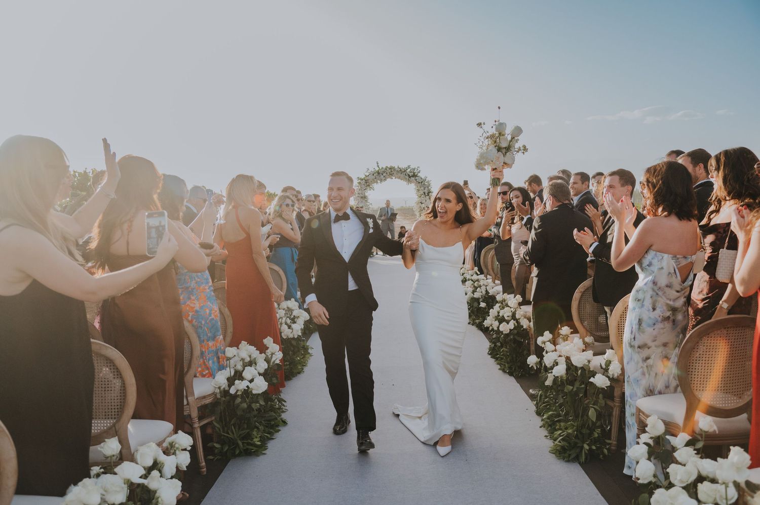 Bride and groom walk down outdoor aisle surrounded by white flowers and cheering guests at a sunny wedding ceremony.