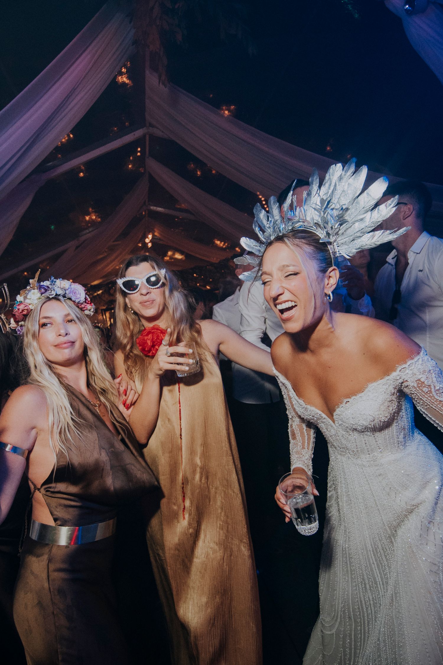 Bride wearing feather crown laughs with two friends in festive attire at wedding reception party.
