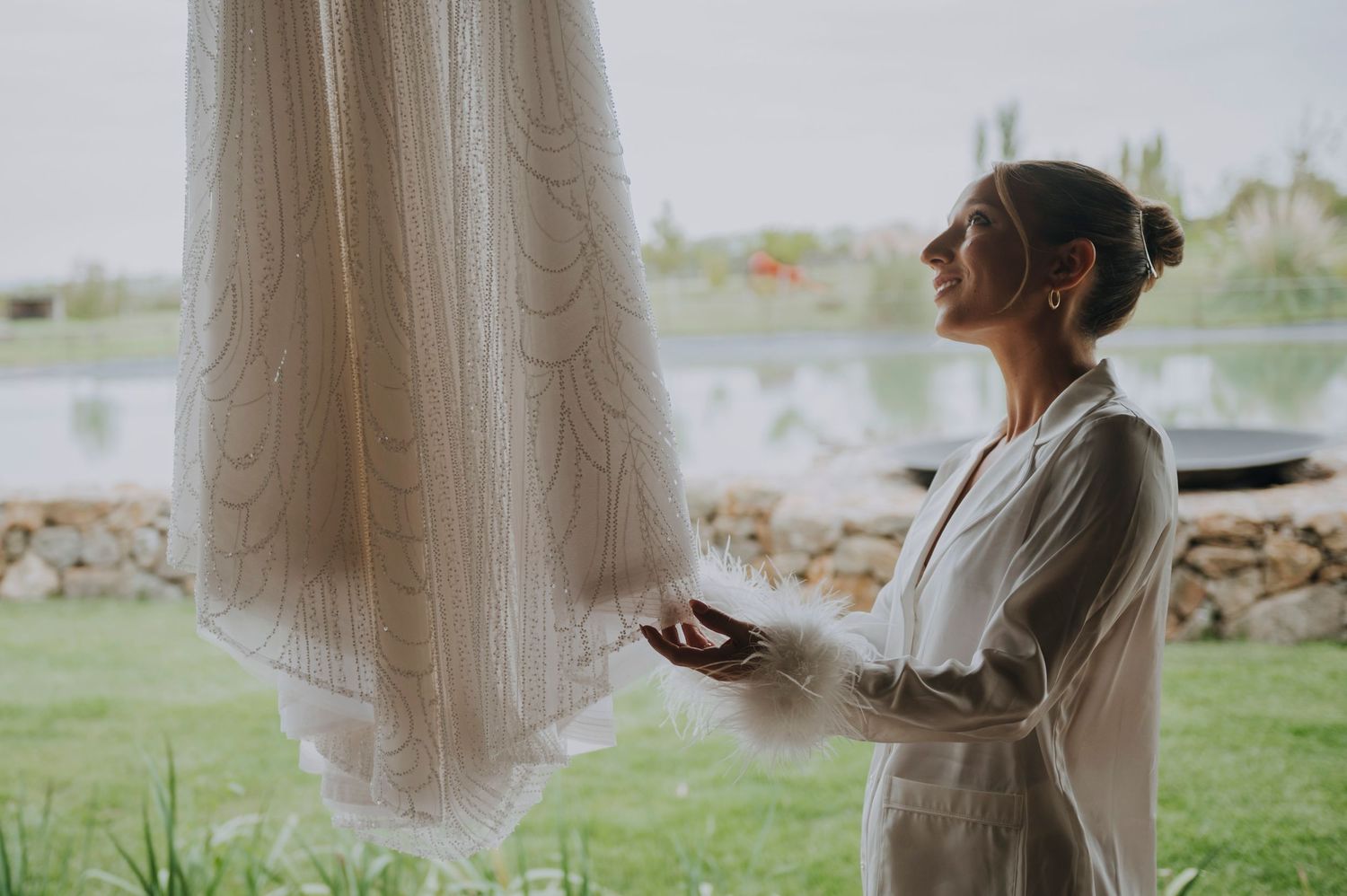 Smiling bride in silk robe admiring her lace wedding dress hanging by a window with scenic lake view.