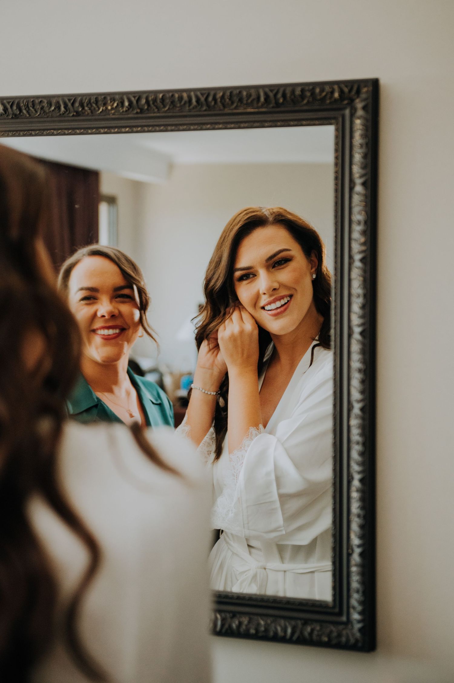 Bride in white robe smiling while putting on earrings reflected in ornate mirror with bridesmaid behind her.