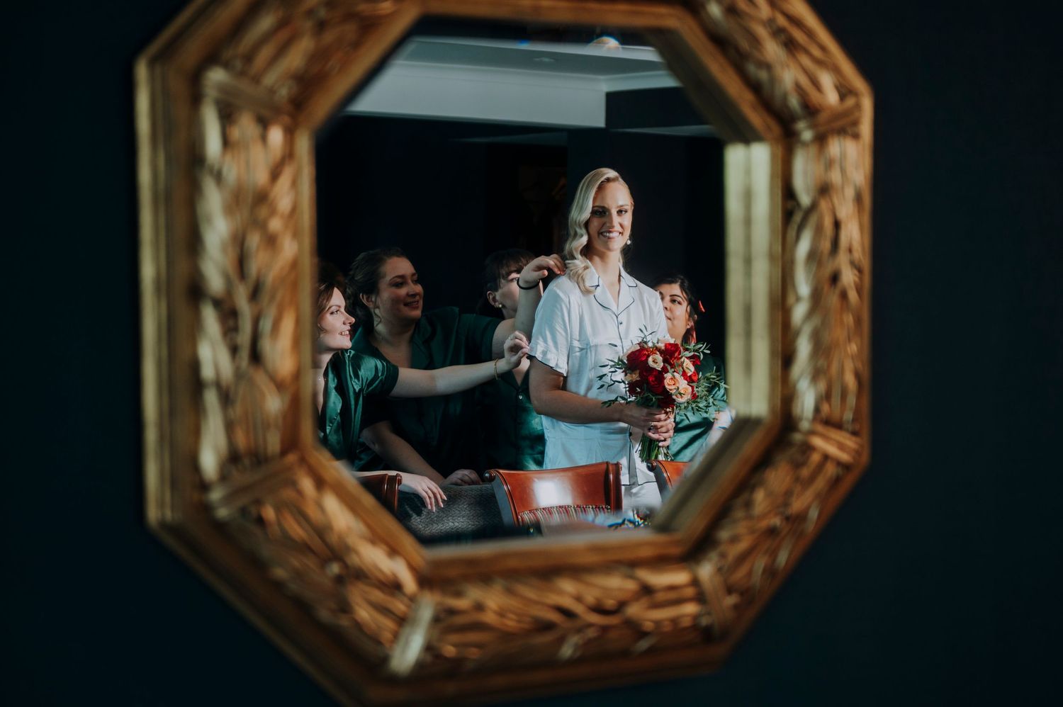 Bride getting ready with bridesmaids helping with hair and jewelry, reflected in ornate gold octagonal mirror.